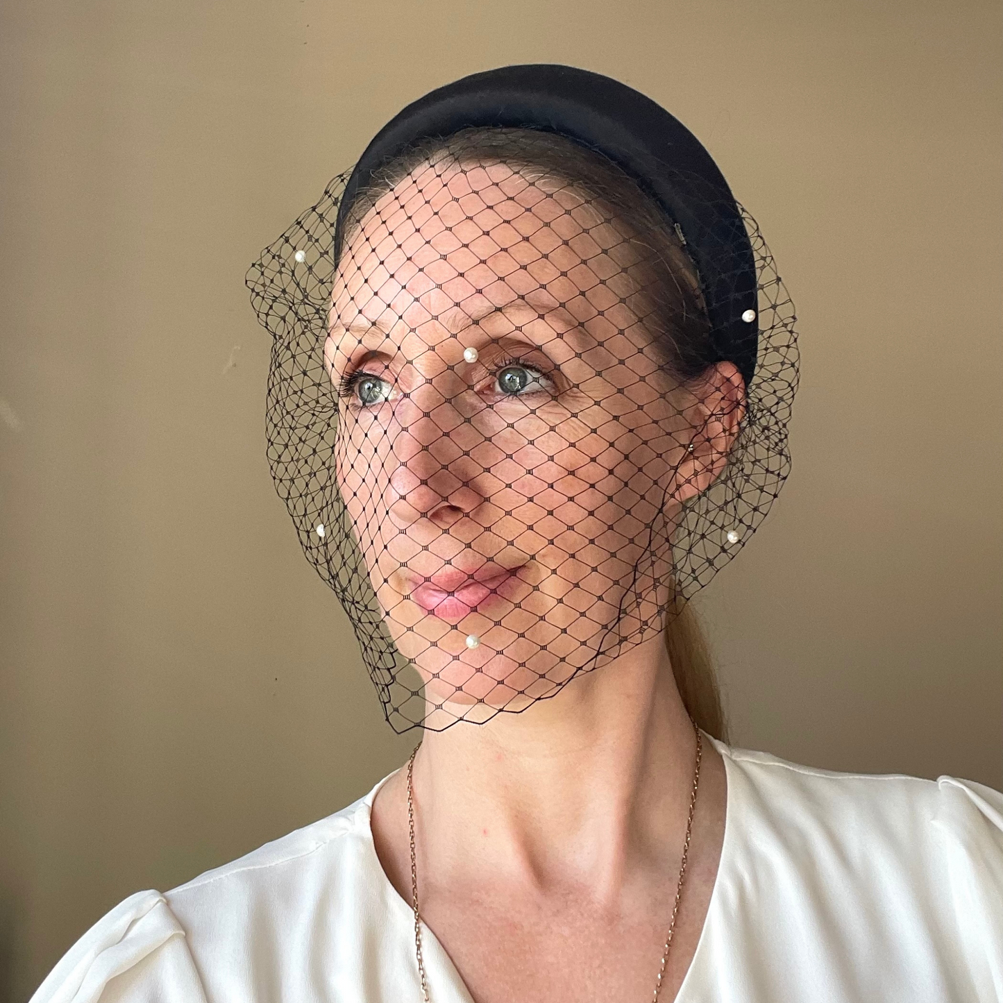 Wearing a black satin covered padded headband with an attached veil that is studded with faux pearls.